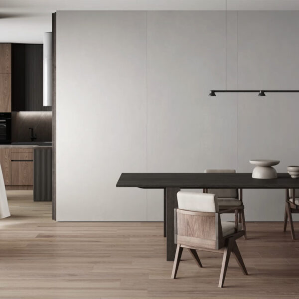 Minimalist dining room with a long dark wooden table, light upholstered chairs, and a gray paneled wall; kitchen visible in the background.
