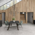 Modern open dining area with a black marble table, green upholstered chairs, a geometric pendant chandelier, wood paneled walls, and a wooden staircase.