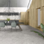 Modern open-concept dining area with grey marble floors, glass table, green upholstered chairs, and wooden wall panels.