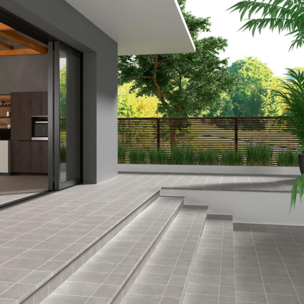 Modern patio with gray tiled steps leading to a sliding glass door into a kitchen, bordered by a wooden fence and lush greenery.