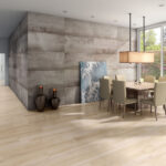 Modern dining room with a long wooden table, beige upholstered chairs, a gray textured wall, and a large rectangular pendant light; art leaning against the wall and two vases on the floor.