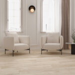 Two cream upholstered armchairs with black legs facing each other in a bright, minimalist living room with light wood flooring and sheer curtains.