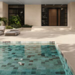 Modern home's beige exterior with a wooden front door, a tiled pool in foreground, and beige lounge chairs beside tropical plants.