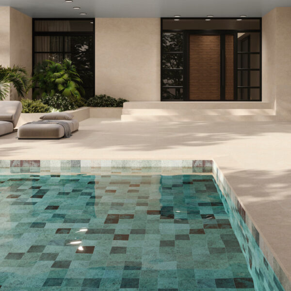 Modern home's beige exterior with a wooden front door, a tiled pool in foreground, and beige lounge chairs beside tropical plants.