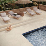Poolside lounge area with four gray cushioned sunbeds under wicker umbrellas along a beige stone deck and planters nearby