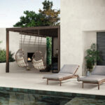 Outdoor lounging area with two hanging wicker chairs under a wooden pergola, by a pool and beige stucco wall with potted plants nearby.