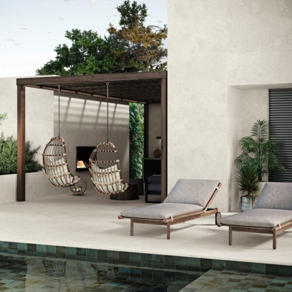 Outdoor lounging area with two hanging wicker chairs under a wooden pergola, by a pool and beige stucco wall with potted plants nearby.