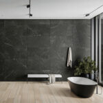 Contemporary bathroom with dark tiled wall, a black freestanding tub, a light wood floor, and a bench with a towel beside a potted plant near a large window.