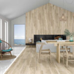 Bright coastal living room with a tall wood-paneled wall, a built-in fireplace, and a wooden dining table; large window shows the sea outside.