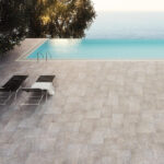 Outdoor pool deck with two black lounge chairs and a white towel, overlooking the sea and a sunlit tree to the left.