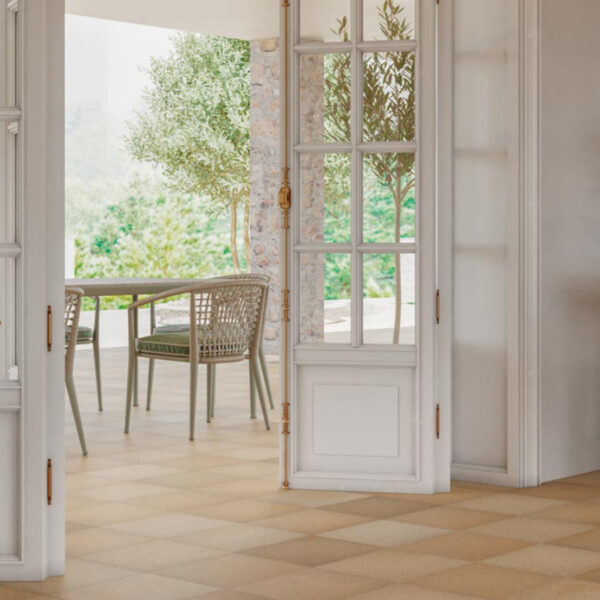 Open white-framed glass doors lead to a sunlit outdoor patio with wicker chairs and a table amid greenery.