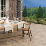 Outdoor dining table set on a stone patio with a pitcher, bowls of fruit, and cups, overlooking a forested landscape.