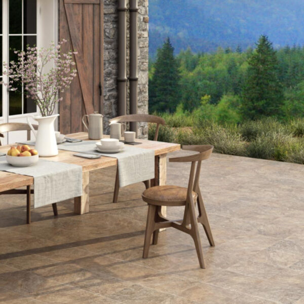 Outdoor dining table set on a stone patio with a pitcher, bowls of fruit, and cups, overlooking a forested landscape.