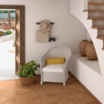Bright entry alcove with white wicker chair and yellow cushion, a potted plant, wall hooks holding hat, bag, and binoculars, and an open wooden door to a sunny patio.