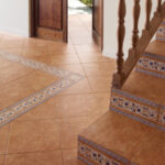 Interior view of terracotta floor tiles with decorative patterned border tiles near a wooden staircase railing and an open doorway to the outdoors.