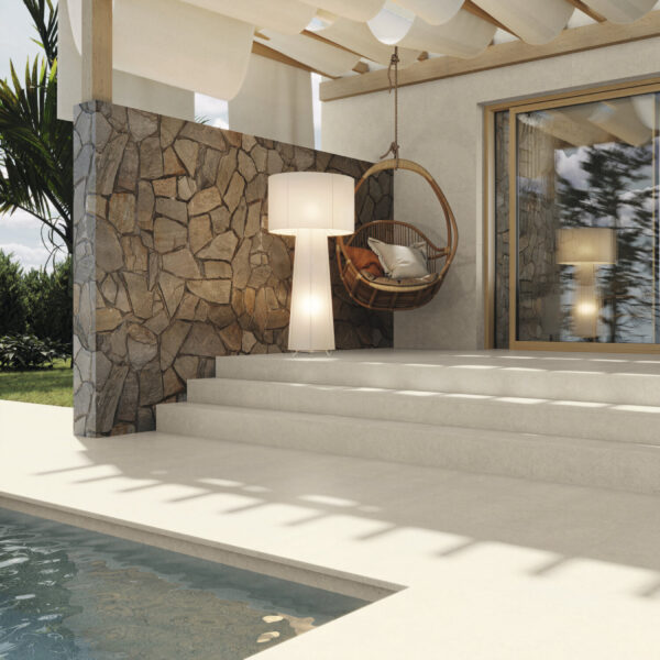 Outdoor patio with a stone wall, a circular hanging wicker chair with cushions, a tall white floor lamp, and concrete steps by a pool.