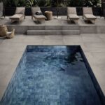 Modern outdoor pool area: gray stone deck, four beige lounge chairs, wicker baskets, and a rectangular blue-tiled pool in the foreground.