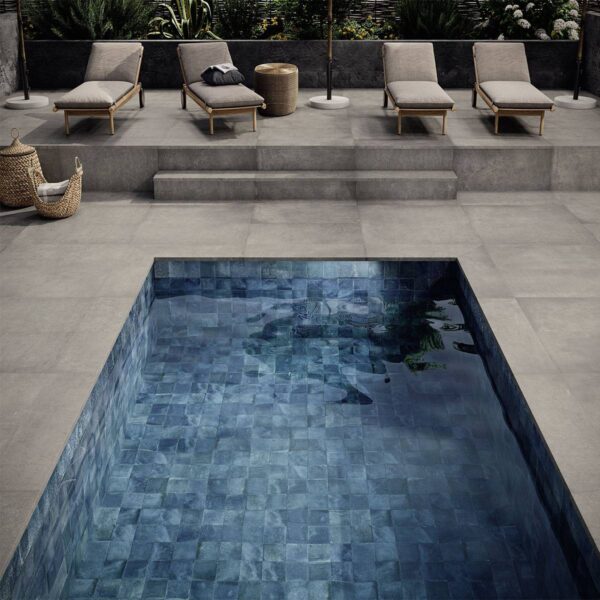 Modern outdoor pool area: gray stone deck, four beige lounge chairs, wicker baskets, and a rectangular blue-tiled pool in the foreground.