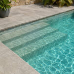 Pool with stone-tiled steps descending into turquoise water, surrounded by a gray concrete deck and tropical plants in the background.