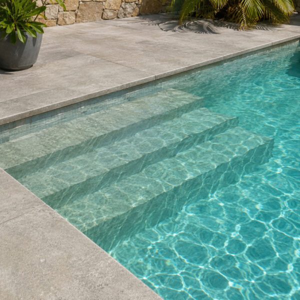 Pool with stone-tiled steps descending into turquoise water, surrounded by a gray concrete deck and tropical plants in the background.